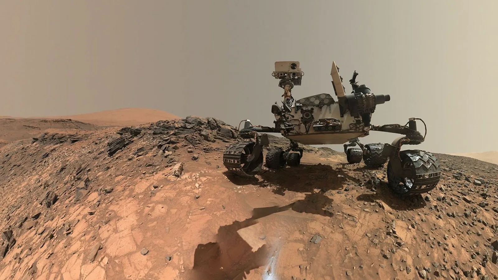 Un rover sobre la superficie marciana anaranjada y ondulada, bajo un cielo gris plano, rodeado de bloques de roca.